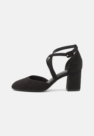 Chaussure à talon bloc en daim noir à bout fermé avec des brides croisées autour de la cheville et une boucle latérale sur un fond blanc.