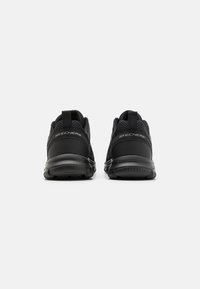 Black athletic shoes with a textured mesh upper, padded collar, pull tab, and cushioned sole. Logo displayed on the heel.