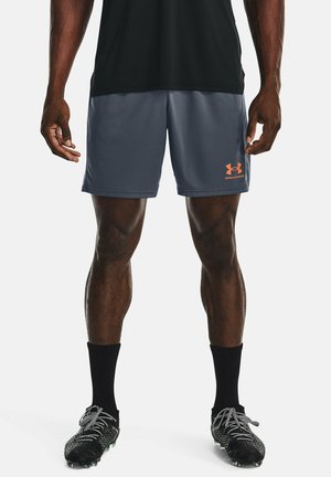 Mann trägt dunkelgraue Under Armour Sportshorts, schwarze Kniesocken und schwarze Fußballschuhe mit weißen Schnürsenkeln, steht vor weißem Hintergrund.