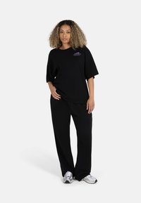 Svart oversized t-skjorte med lilla grafikkdetalj, kombinert med svarte, vide bukser og sølvfargede sneakers. Enkelt, uformelt design.