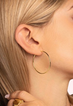 Goldene Kreolen, die im Ohr platziert sind, mit einer glatten, metallischen Oberfläche und einem kreisförmigen Design. Die Haare sind glatt und blond.
