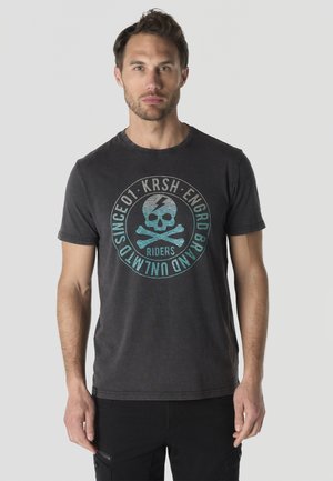 Hombre con el pelo corto que lleva una camiseta negra con un gráfico de calavera y huesos cruzados y texto alrededor del diseño, de pie contra un fondo liso.