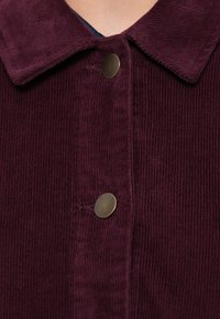 Giacca in velluto a coste color bordeaux con colletto, caratterizzata da due bottoni rotondi in bronzo e un tessuto strutturato a coste. Vista ravvicinata dell'area di chiusura.