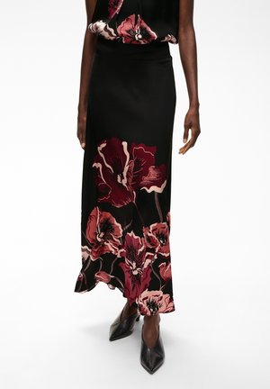 Femme portant une jupe noire avec de grandes impressions florales rouge foncé associée à un haut fleuri assorti et des chaussures noires à bout pointu.