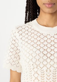 Maglione bianco lavorato a maglia con un motivo a rombi, scollo a costine e maniche corte con bordi ondulati. Tessuto leggero e dalla texture piacevole.
