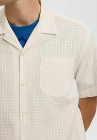 Korte mouwen met kraag in roomkleur met een gestructureerd ruitpatroon, enkele borstzak en transparante knopen, gedragen over een blauwe shirt.
