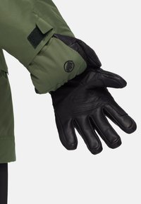 Grüne Außenlage mit schwarzen Lederhandschuhen. Verfügt über einen verstellbaren Bund und eine strukturierte Handfläche für besseren Grip. Markenlogo am Bündchen sichtbar.