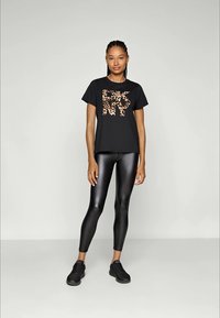 Junge Frau trägt ein schwarzes DKNY T-Shirt mit Leopardenmuster, schwarze Leggings aus Kunstleder und schwarze Sneakers, steht vor einem schlichten Hintergrund.