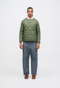 Jachetă verde cu quiltare, cu guler rotund și închidere prin capse. Pantaloni largi din denim albastru și pantofi tip bărci maro completează ținuta.