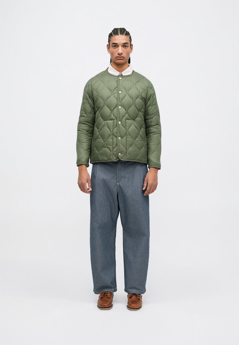 Jachetă verde cu quiltare, cu guler rotund și închidere prin capse. Pantaloni largi din denim albastru și pantofi tip bărci maro completează ținuta.