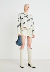 Hvit strikkegenser med svart tekstmønster, beige mini skjørt med belte, lyse hæler, og en blå stoffhåndveske.