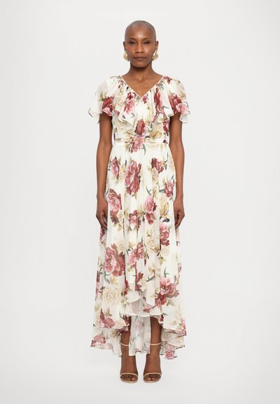 Blumenkleid in mittellanger Form aus durchsichtigem Material mit Rüscheärmeln. Es zeigt Farbnuancen von Creme, Pink und Grün mit einem asymmetrischen Saum. Trägt Riemchensandalen.