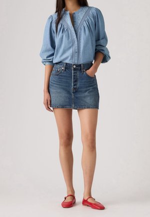 Kvinde iført en lyseblå bluse med lange ærmer, denim mini nederdel og røde flade sko, stående med den ene hånd i lommen.