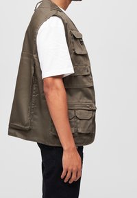 Olivgrüne ärmellose Weste mit mehreren Taschen, gefertigt aus einem robusten Stoff, ausgestattet mit Reißverschluss und Druckknöpfen.