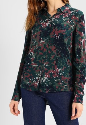 Woman wearing a long-sleeve button-up shirt with green, black, red, and white abstract camouflage print and dark blue jeans.