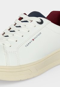 Άσπρο αθλητικό παπούτσι Tommy Hilfiger με άσπρα κορδόνια, μπεζ σόλα, ναυτικό και κόκκινο επενδυμένο κολάρο και λογότυπο της μάρκας στο πλάι.