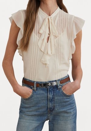 Kvinde iført en ærmeløs elfenbensfarvet bluse med flæser på skuldrene og bindebånd foran, stoppet ned i blå jeans med et brunt læderbælte.