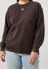 Felpa marrone tie-dye con vestibilità ampia, polsini a coste e finitura testurizzata. Abbinata a denim blu chiaro.
