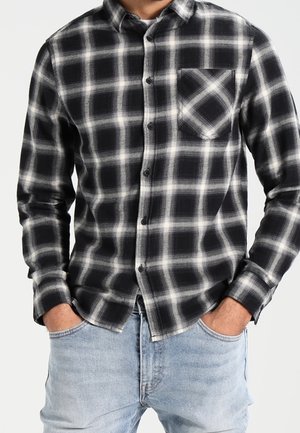 Mand iført en sort og hvid ternet skjorte med knapper og brystlomme, hænderne i lommerne, kombineret med lyseblå jeans.