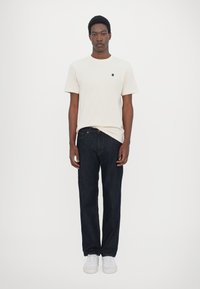 Kortsleeved beige t-skjorte med en svart logo, sammen med mørkeblå jeans og hvite joggesko, alt mot en enkel hvit bakgrunn.