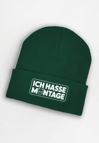Green knitted beanie featuring a white rectangular patch with "ICH HASSE MONTAGE" text and a cat graphic. Ribbed texture throughout.