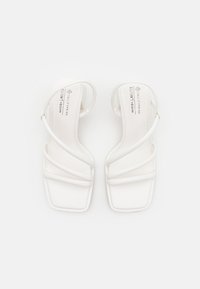 Sandales véganes blanches avec un bout carré, un design minimaliste à lanières et des brides de cheville réglables. Texture lisse et mate.