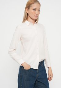 Svetlo roza srajca na gumb s ovratnikom, dolgimi rokavi in rahlo prilegajočo obliko, kombinirana z modrimi jeans hlačami. Gladka tekstura tkanine.