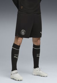 Ben klædt i sorte Manchester City fodboldshorts, sorte knæhøje sokker med Puma-logo og hvide fodboldstøvler foran en grå baggrund.