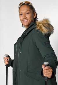 Veste isolée verte avec une capuche en fourrure, fermeture éclair à l'avant et boutons-pression. Deux poches avant et une texture lisse sont visibles.