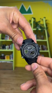 Zwarte Lacoste-horloge met een ronde kast, digitale en analoge display, groene accentdetails en een textuur rubberen band, vastgehouden in een hand.