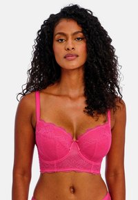 Růžová krajková podprsenka s kosticemi, vlnkovanými okraji a mašlí uprostřed. Ramínka jsou nastavitelná pro přizpůsobenou velikost.