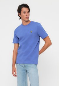 T-shirt bleu à manches courtes en coton doux, arborant un petit logo d'oiseau jaune brodé sur la poitrine gauche et un col rond.