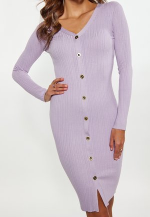 Woman wearing a fitted, ribbed lilac dress with long sleeves and front buttons, posing with one hand on her hip against a plain background.