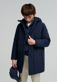 Marineblå hættejakke med lynlås, sidelommer, og lagdelt over en blå striktrøje. Lys tan bukser og en blå cap i hånden.