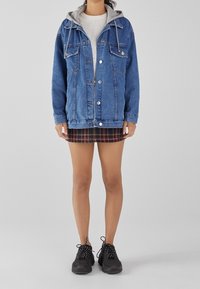 Spijkerjack met een grijze capuchon, twee borstzakken en knoopsluiting, gecombineerd met een geruite minirok en zwarte sneakers. Effen achtergrond.