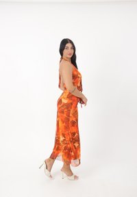 Maxi jurk in oranje met bloemenpatronen, halternek en zijsplitsingen. Gecombineerd met witte hoge muiltjes met een gestructureerd ontwerp.