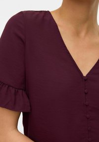 Bordeaux blouse met een V-hals, rimpelige korte mouwen en een rij knoopdetails aan de voorkant. Zachte, gladde stoftextuur.