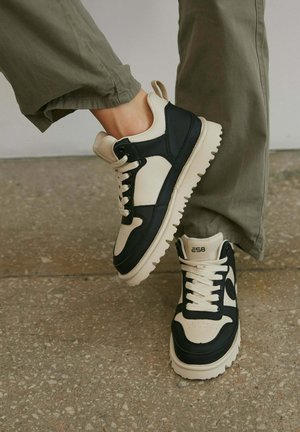 Sneaker z beżową skórzaną cholewką, granatowymi akcentami i masywną białą gumową podeszwą. Cechy obejmują okrągłe sznurowadła i logo na pięcie.