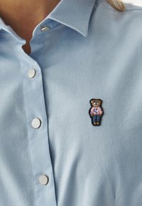 Lichtblauwe, knooploze shirt met een klein geborduurd beertje in een roze shirt en blauwe spijkerbroek op de linkerborst. Soepele stoftextuur.