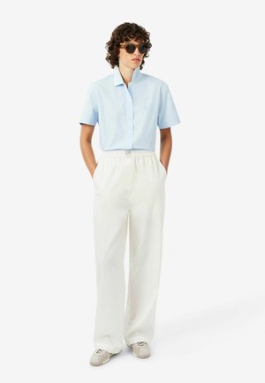Persoon draagt lichtblauw kort mouw overhemd met knopen, losse witte broek, beige sneakers en donkere zonnebril, staat met handen in de zakken.