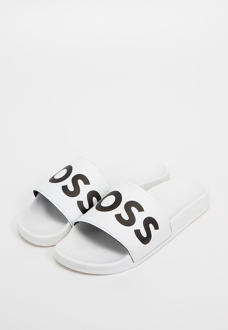 Des sandales blanches à glissière avec une large sangle arborant des lettres noires audacieuses. Texture lisse et design simple, avec une semelle texturée pour une meilleure adhérence.