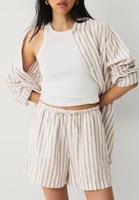 Pantalones cortos de lino a rayas y una camisa oversized a juego en tonos beige y blanco apagado. La camiseta de tirantes es completamente blanca.