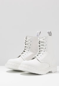 Stivali da combattimento in pelle bianca con punta rotonda, texture liscia e lacci bianchi. Presentano un anello sul tallone e una suola bianca abrasiva.