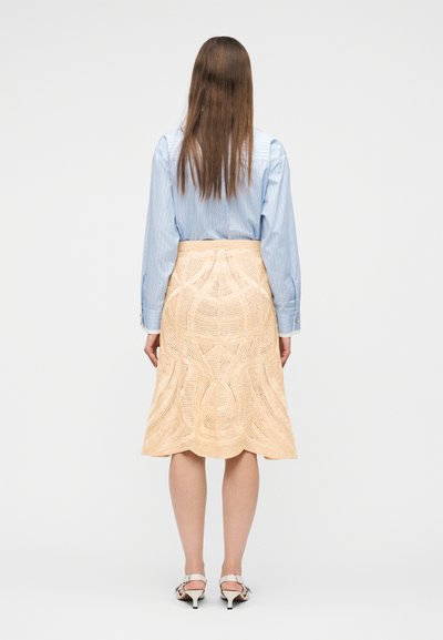 Femme debout de dos portant une chemise à rayures bleues, une jupe beige texturée longueur genou et des sandales blanches à talons sur un fond blanc.