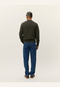 Olivgrön stickad tröja med ribbade muddar och nederkant, kombinerad med blå denimjeans. Utsikt från baksidan, med en basebollkeps.