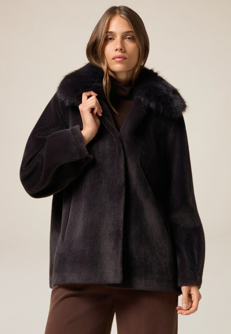 Abrigo de piel oscura con un cuello de pelo sintético, diseño oversized, mangas largas y una textura suave, exhibido sobre un fondo neutro.