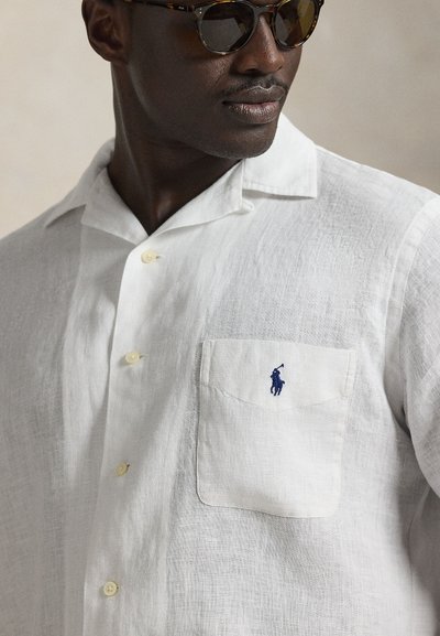 Balta linu īsroku krekls ar krūšu kabatu un tumši zilu izšūtu logotipu. Ir gaišas pogas un relaksēts apkakles dizains.