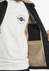 Black plaid jacket with a beige fleece lining. White t-shirt features a round embroidered logo in blue and black. Open jacket reveals interior pocket.