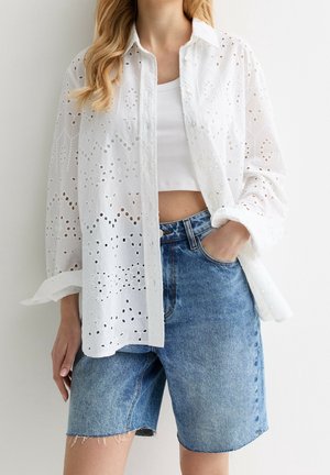 Kvinde iført hvid øjlet knappet skjorte over hvid cropped top og løstsiddende denimshorts, med hånden i lommen, foran en ensfarvet baggrund.