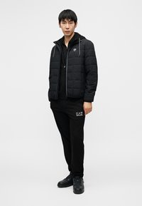 Μαύρο καπιτονέ μπουφάν με φερμουάρ μπροστά, συνδυασμένο με μαύρο φούτερ με κουκούλα και φόρμες. Το σύνολο διαθέτει διακριτικό λογότυπο στα παντελόνια και τα παπούτσια.